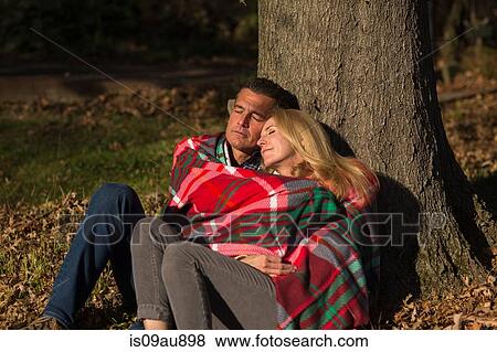 Stock Photo - Mature couple sitting against tree trunk wrapped in blanket at night. Fotosearch