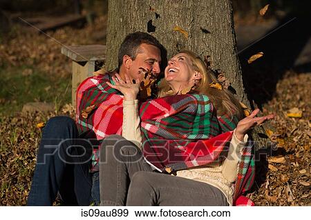 Stock Photo - Mature couple sitting against tree trunk throwing autumn leaves at night. Fotosearch