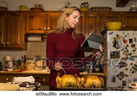 Picture - Mature woman using digital tablet to check roast chicken temperature in kitchen. Fotosearch
