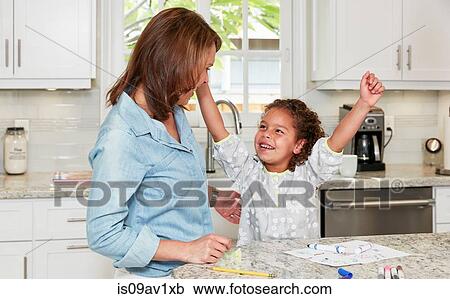 Stock Image - Mother and daughter at kitchen counter drawing pictures, arms raised triumphantly, smiling. Fotosearch