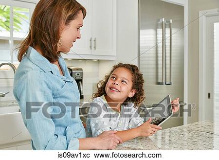 Mother and daughter at kitchen counter using digital tablet, looking up smiling View Large Photo Image Stock Image - Mother and daughter at kitchen counter using digital tablet, looking up smiling. Fotosearch