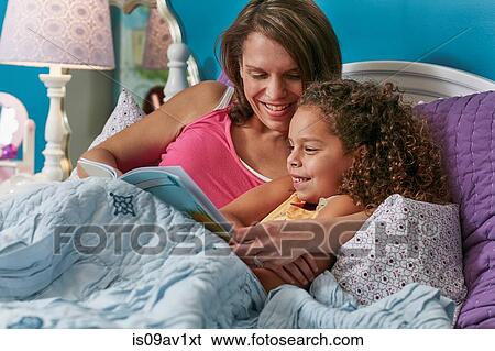 Stock Photograph - Mother and daughter cuddling in bed reading book smiling. Fotosearch