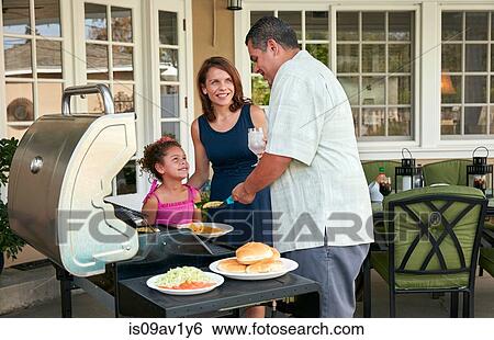 Stock Photograph - Mother and girl on patio holding plate being served barbeque food by father. Fotosearch