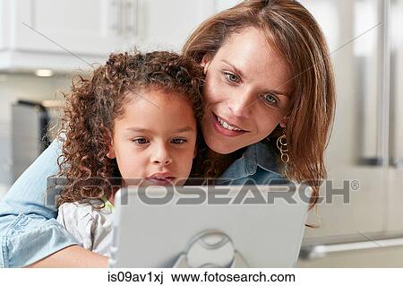 Stock Image - Mother with arm around daughter using digital tablet looking down smiling. Fotosearch