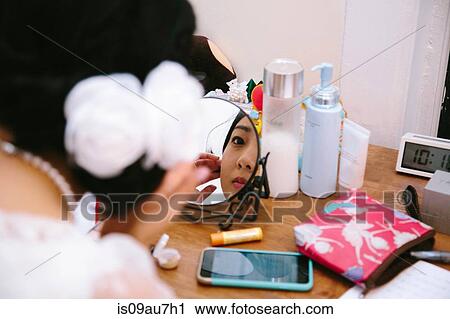 Stock Image - Over the shoulder view of young woman at dressing table applying makeup in mirror. Fotosearch