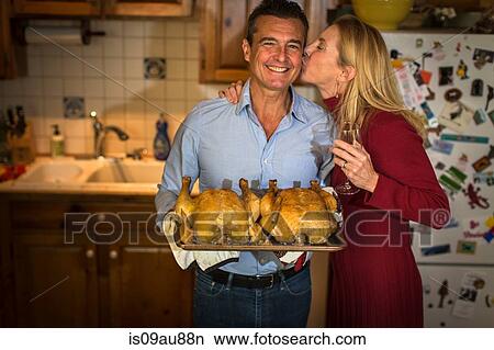 Picture - Portrait of mature couple holding roast chickens in kitchen. Fotosearch