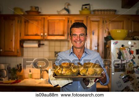 Picture - Portrait of mature man holding roast chickens in kitchen. Fotosearch