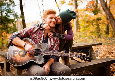 Stock Image - Romantic young couple playing guitar on picnic bench in autumn forest. Fotosearch