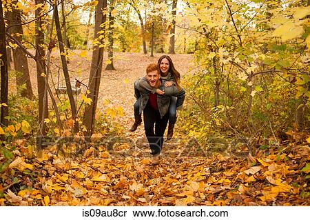 Stock Photography - Romantic young man giving girlfriend a piggy back through autumn forest. Fotosearch