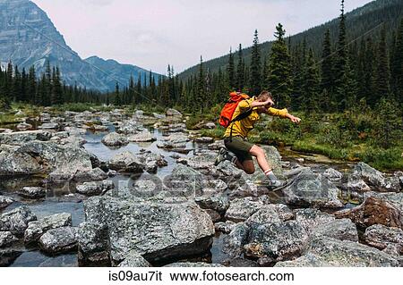 Stock Photograph - Side view of mid adult man carrying backpack jumping over rocky riverbed, Moraine lake, Banff National Park, Alberta Canada. Fotosearch