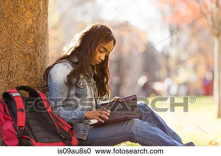 Stock Image - Side view of young woman sitting leaning against tree looking down at digital tablet. Fotosearch