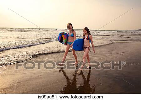 Stock Image - Teenage girl and sister carrying surfboards on beach, Goa, India. Fotosearch