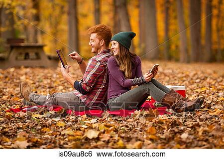 Stock Photograph - Young couple back to back using digital tablet and smartphone in forest. Fotosearch