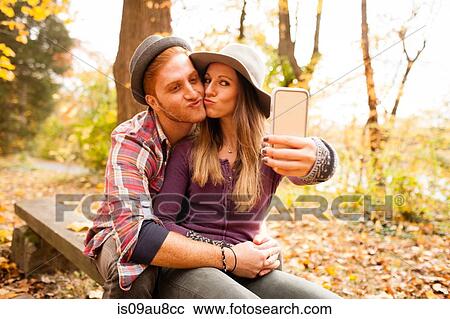 Stock Image - Young couple on bench taking smartphone selfie in autumn forest. Fotosearch