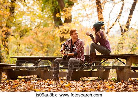 Stock Photo - Young couple playing guitar on picnic benches in autumn forest. Fotosearch