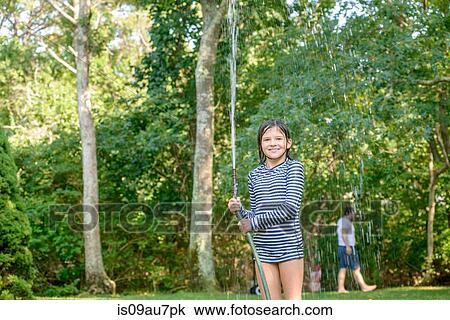 Stock Image - Young girl in garden, holding garden hose. Fotosearch