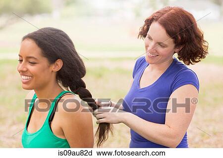 Stock Image - Young woman behind friend plaiting hair smiling. Fotosearch