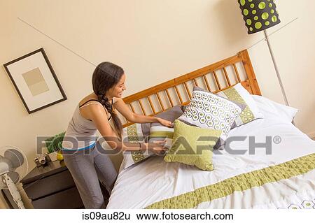 Stock Photograph - Young woman in bedroom placing cushions on bed. Fotosearch