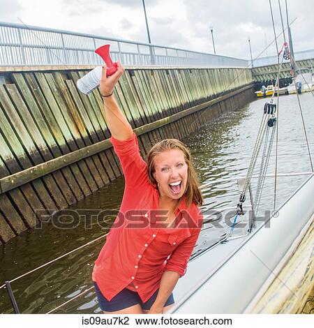Stock Image - Young woman on sailboat in marina arm raised holding air horn, looking at camera open mouthed smiling. Fotosearch