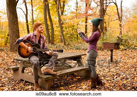 Stock Photo - Young woman photographing guitar playing boyfriend in autumn forest. Fotosearch