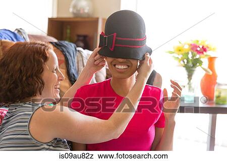 Stock Photo - Young woman putting hat on smiling friends head, covering eyes. Fotosearch