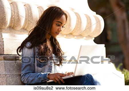 Stock Image - Young woman sitting leaning against stone carving looking down using laptop computer. Fotosearch