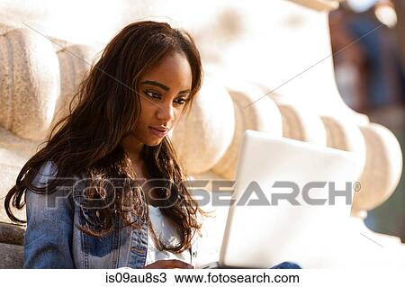 Stock Image - Young woman sitting leaning against stone carving looking down using laptop computer. Fotosearch
