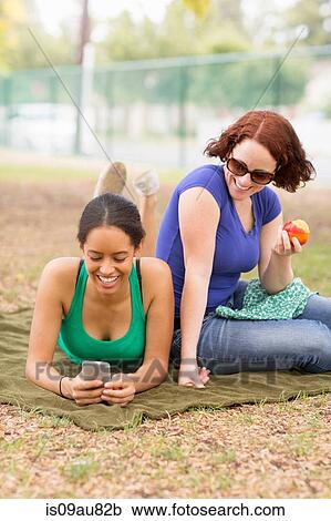 Stock Image - Young women on picnic blanket using smartphone looking down smiling. Fotosearch