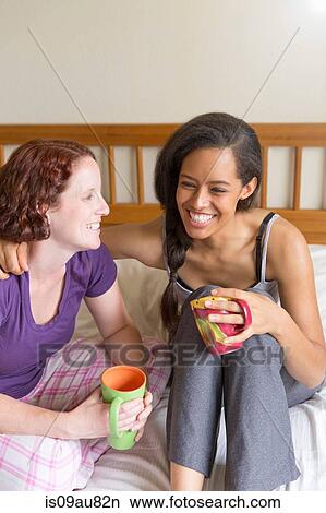 Picture - Young women sitting on bed, arm around friend, holding cup smiling. Fotosearch