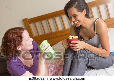 Young women sitting on bed holding cups, chatting, smiling View Large Photo Image Picture - Young women sitting on bed holding cups, chatting, smiling. Fotosearch