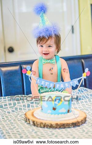 Stock Image - Baby boy wearing party hat with first birthday cake at table. Fotosearch