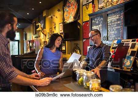 Stock Image - Barman reading menu with young couple at public house counter. Fotosearch