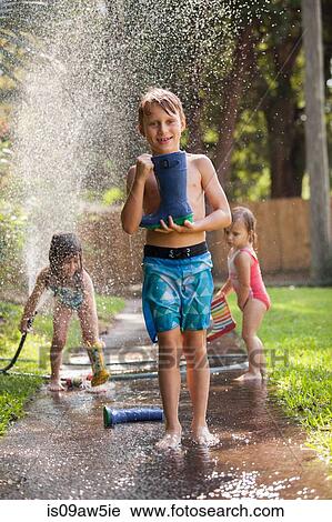 Stock Image - Boy carrying welly on sidewalk, girls playing with water hose in background. Fotosearch