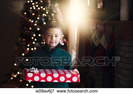 Boy in front of christmas tree holding gift looking at camera smiling View Large Photo Image Stock Photo - Boy in front of christmas tree holding gift looking at camera smiling. Fotosearch