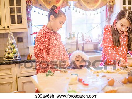 Stock Image - Boy in kitchen wearing santa hat making cookies with sisters peeking over kitchen counter open mouthed. Fotosearch
