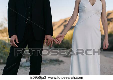 Stock Image - Bride and groom, in arid landscape, touching hands, mid section. Fotosearch