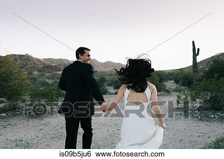 Stock Photograph - Bride and groom in arid landscape, holding hands, running, rear view. Fotosearch