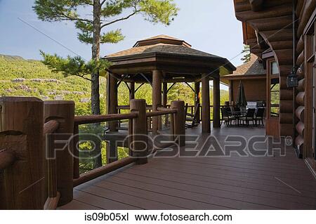 Stock Image - Brown gazebo on wooden deck with tempered glass and log railings at back of luxurious cottage style log home in summer, Quebec, Canada. Fotosearch