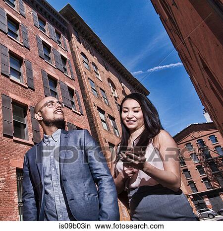 Business man and woman walking outdoors, using smartphone, low angle view View Large Photo Image Picture - Business man and woman walking outdoors, using smartphone, low angle view. Fotosearch