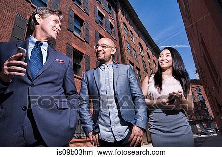 Picture - Business men and women walking outdoors, holding smartphone, smiling, low angle view. Fotosearch