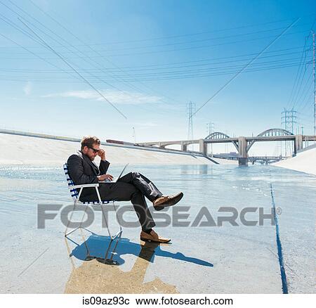Stock Image - Businessman in deckchair on Los Angeles river using smartphone and laptop, California, USA. Fotosearch