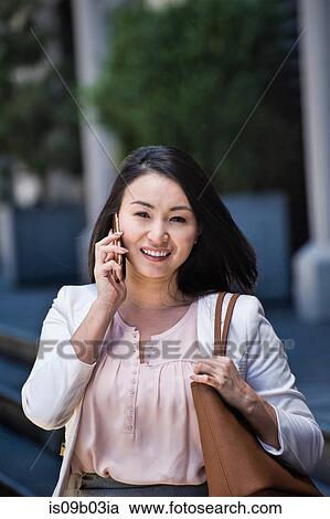 Stock Image - Businesswoman walking outdoors, using smartphone, smiling. Fotosearch