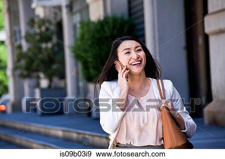 Stock Photo - Businesswoman walking outdoors, using smartphone, smiling. Fotosearch
