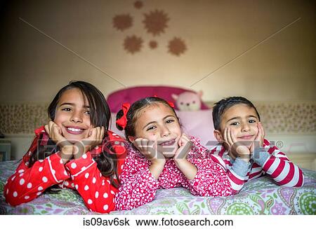 Children wearing pyjamas lying on fronts on bed chin in hands looking at camera smiling View Large Photo Image Stock Image - Children wearing pyjamas lying on fronts on bed chin in hands looking at camera smiling. Fotosearch