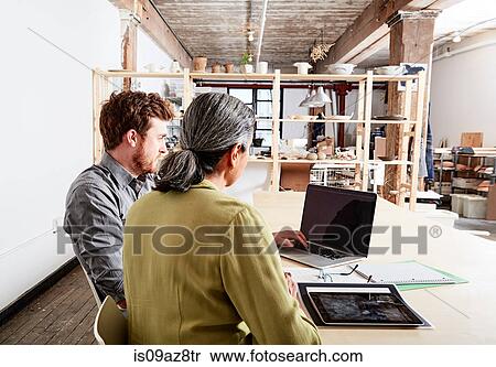 Stock Photography - Colleagues at desk in workshop looking at laptop computer. Fotosearch