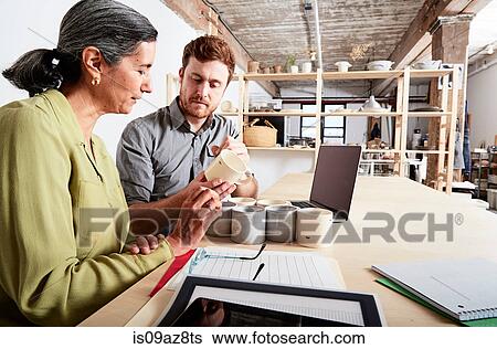 Colleagues at desk in workshop discussing pottery mugs View Large Photo Image Stock Photograph - Colleagues at desk in workshop discussing pottery mugs. Fotosearch