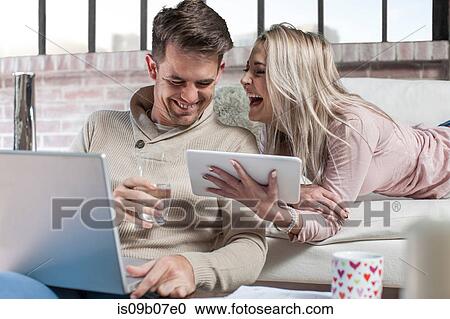 Stock Image - Couple at home, man using laptop, woman holding digital tablet, laughing. Fotosearch