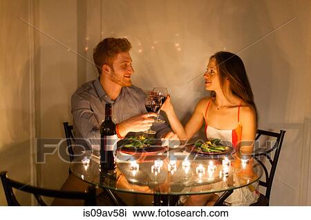 Stock Image - Couple at table face to face enjoying candlelit meal, making a toast, smiling. Fotosearch