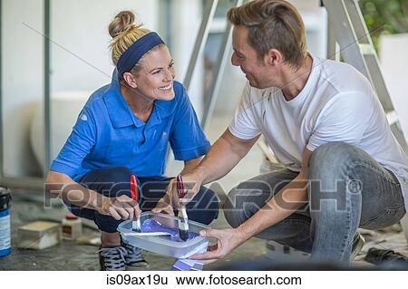 Stock Photograph - Couple crouching dipping paintbrushes in plastic container face to face smiling. Fotosearch