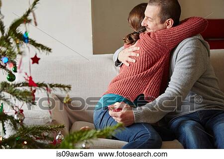 Picture - Couple holding xmas gift hugging on sofa at xmas. Fotosearch